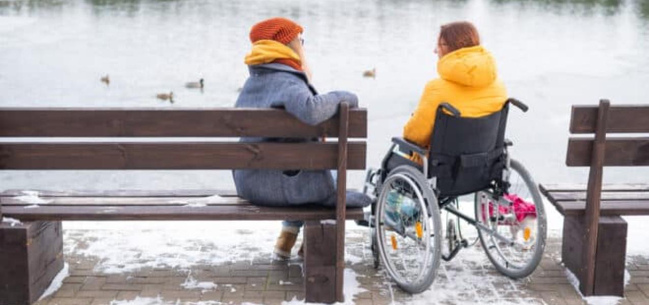 Two people sitting next to each other at lake, one in a wheel chair